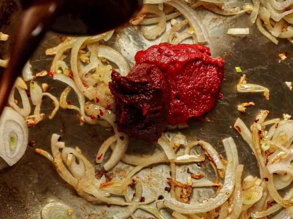 Onions and tomatoes are being cooked in a pan. The shallots are golden brown, and the tomatoes appear as a rich, deep red paste (gochujang and tomato paste), suggesting the beginnings of a sauce or stew.